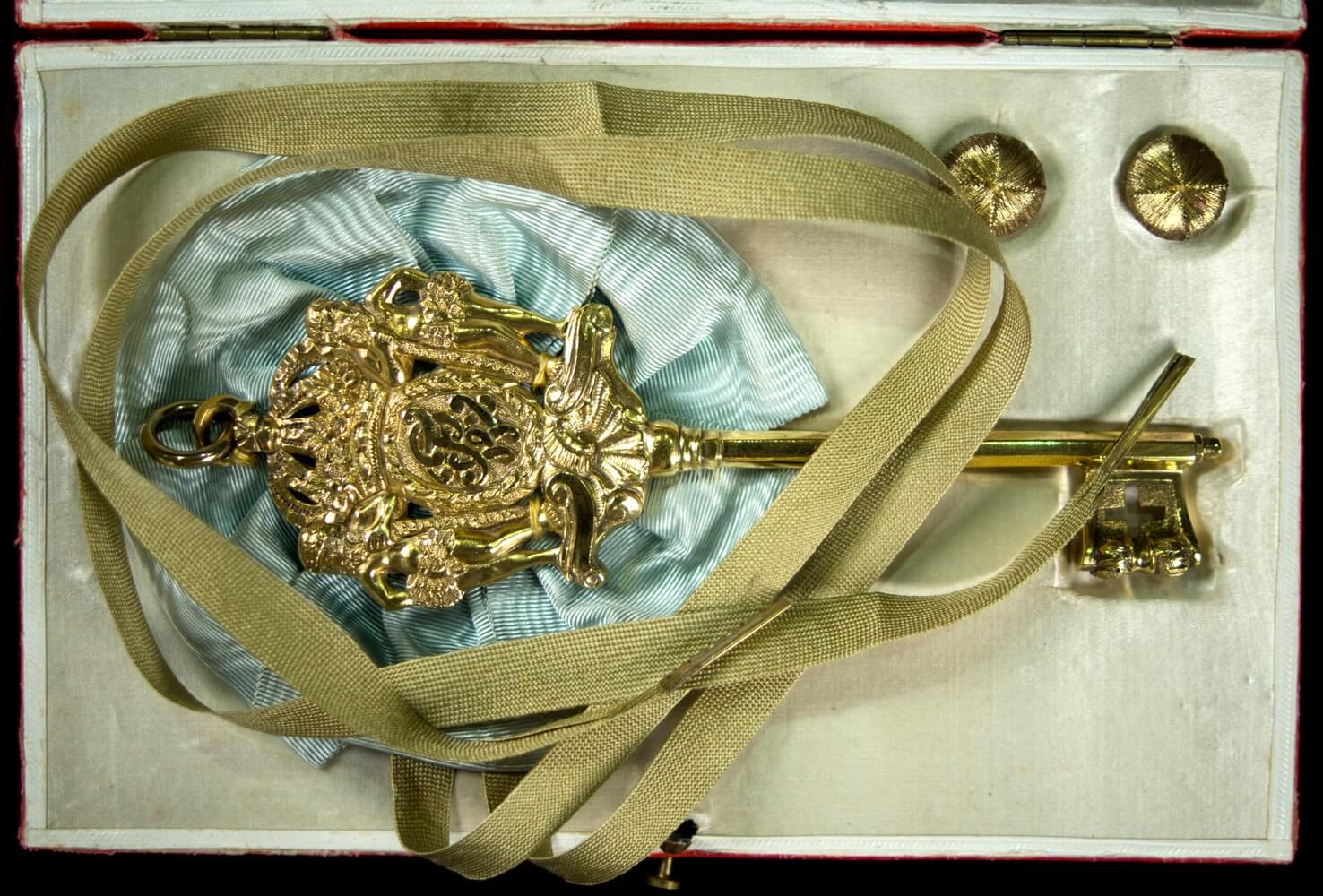 Ein geöffnetes Etui mit einem preußischen Kammerherrenschlüssel, der an einer blauen Seidenrosette mit langen Befestigungsbändern befestigt ist, daneben zwei kleine Befestigungsknöpfe.